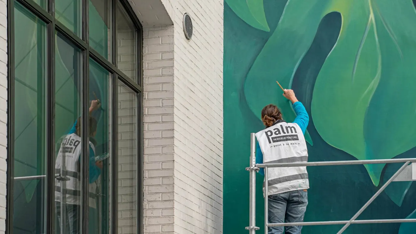 Une artiste peint une fresque aux tons verts sur un mur intérieur.